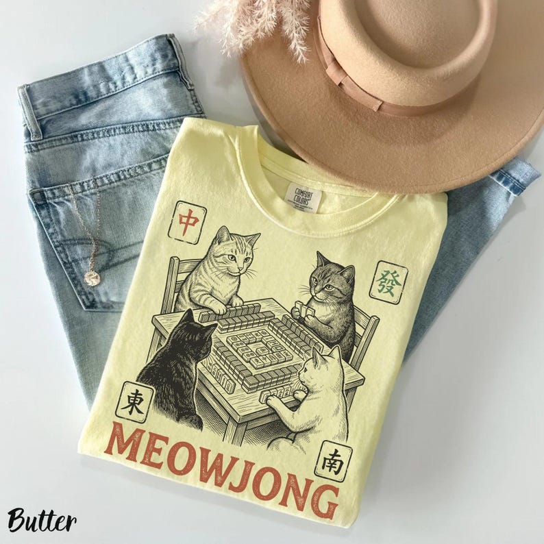 Może przedstawiać: Bladoż&oacute;łty t-shirt z grafiką przedstawiającą cztery koty grające w mahjonga przy stole. Wz&oacute;r zawiera chińskie znaki w kwadratowych ramkach oraz napis MEOWJONG dużymi czerwonymi literami, ułożony na jasnoniebieskich dżinsach.