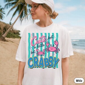 May include: A white short-sleeved t-shirt featuring a graphic print with vertical teal stripes. The design includes two pink crabs and the text "Cute but a little bit CRABBY" in pink and blue lettering with small star and heart accents.