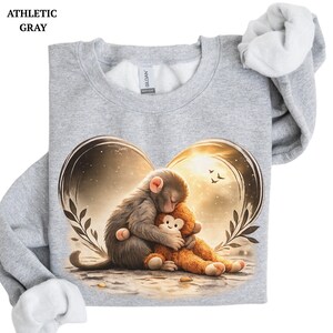 Athletic gray sweatshirt featuring a heart-shaped design with a sleeping monkey hugging a stuffed animal. The graphic includes a warm, golden background with birds and foliage. The sweatshirt has a crew neck and long sleeves.