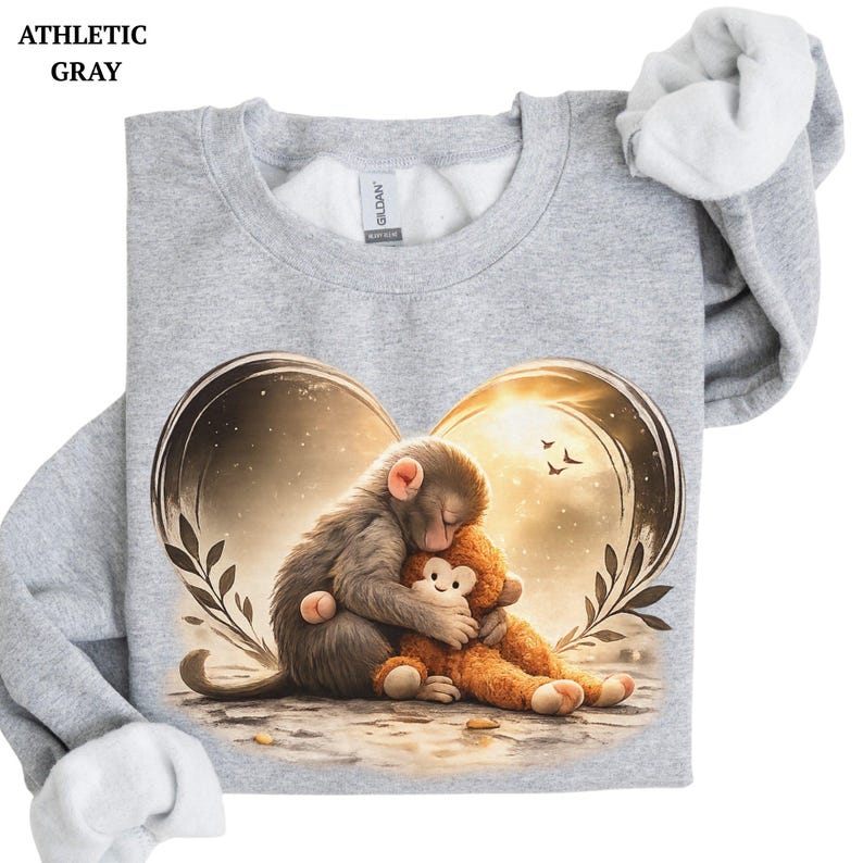 Athletic gray sweatshirt featuring a heart-shaped design with a sleeping monkey hugging a stuffed animal. The graphic includes a warm, golden background with birds and foliage. The sweatshirt has a crew neck and long sleeves.