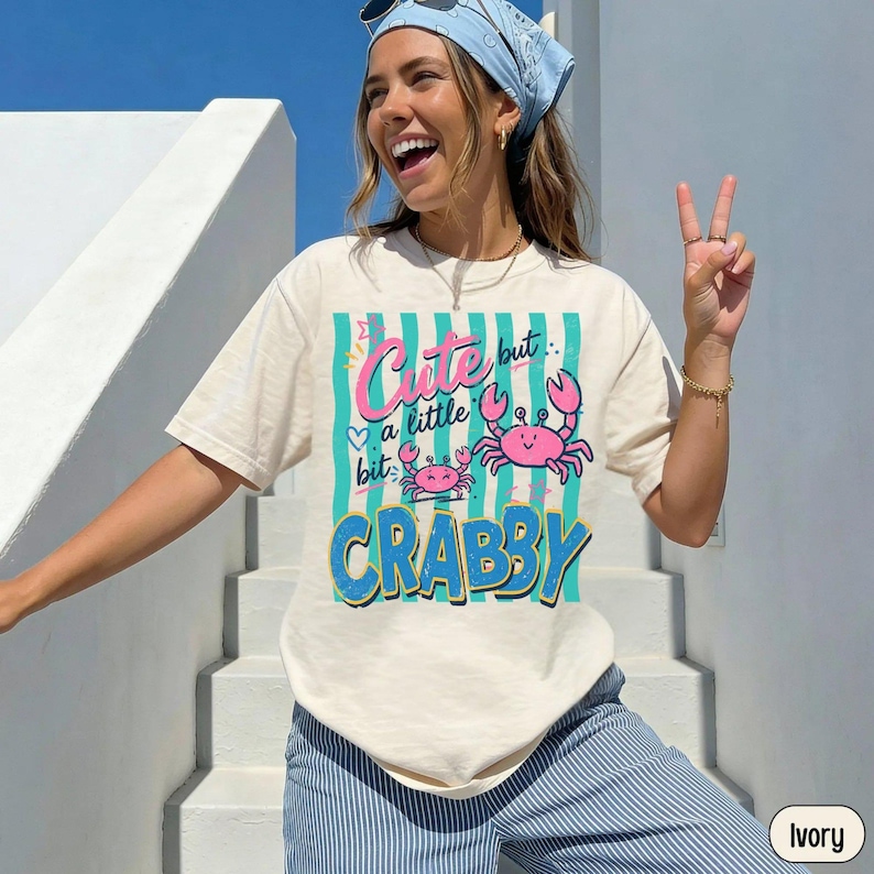 A smiling woman wears an ivory shirt featuring a teal striped graphic with two pink crabs and text reading Cute but a little bit CRABBY.