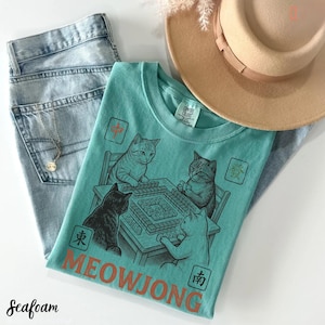 Może przedstawiać: Morski t-shirt z grafiką przedstawiającą cztery koty grające w mahjonga przy stole. Wz&oacute;r zawiera napis MEOWJONG oraz r&oacute;żne chińskie znaki wykonane czerwoną i czarną farbą. Koszulka zestawiona jest z jasnoniebieskimi dżinsami i beżowym kapeluszem.