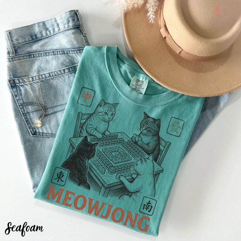 Może przedstawiać: Morski t-shirt z grafiką przedstawiającą cztery koty grające w mahjonga przy stole. Wz&oacute;r zawiera napis MEOWJONG oraz r&oacute;żne chińskie znaki wykonane czerwoną i czarną farbą. Koszulka zestawiona jest z jasnoniebieskimi dżinsami i beżowym kapeluszem.