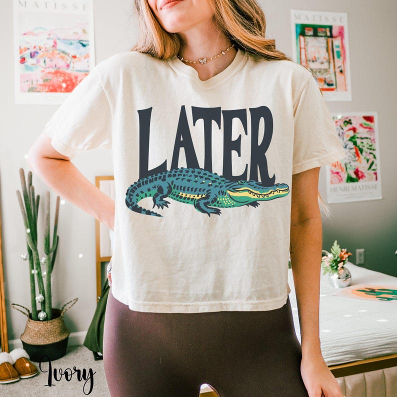 May include: An ivory short-sleeved t-shirt featuring the word LATER printed in large dark gray capital letters above a graphic illustration of a green alligator with yellow accents. The shirt has a crew neckline and a relaxed fit.