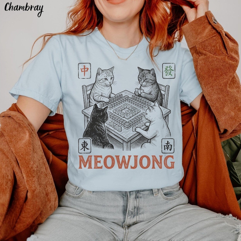 Może przedstawiać: Jasnoniebieski t-shirt z grafiką przedstawiającą cztery koty siedzące przy stole do mahjonga. Wz&oacute;r zawiera chińskie znaki w czterech rogach oraz napis MEOWJONG wydrukowany czerwonobrązową czcionką pod ilustracją stołu.