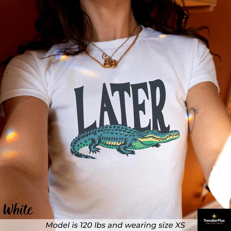 May include: A white short-sleeved t-shirt featuring the word LATER printed in large dark gray capital letters above a graphic illustration of a green and teal alligator. The design is centered on the chest of the garment.