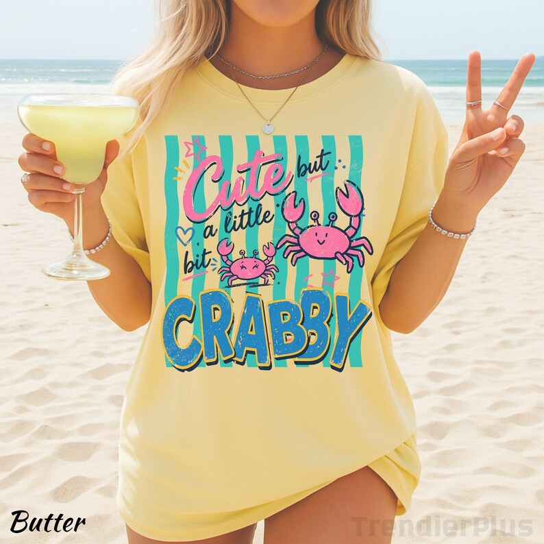 May include: A pale yellow t-shirt featuring a graphic print with teal vertical stripes and two pink crabs. The text reads Cute but a little bit CRABBY in pink and blue lettering. The design includes small heart and star accents.