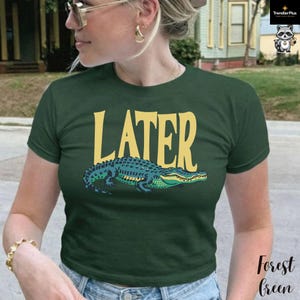 May include: A forest green short-sleeved t-shirt featuring the word LATER printed in large yellow capital letters above a graphic illustration of a teal and green alligator. The shirt has a crew neckline and a standard fit design.