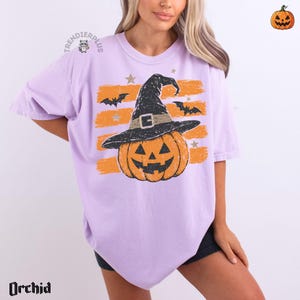 Puede incluir: Una camiseta de color morado claro con un gr&aacute;fico de una calabaza naranja que lleva un sombrero de bruja negro. El dise&ntilde;o incluye trazos de pincel naranjas horizontales, murci&eacute;lagos negros y estrellas peque&ntilde;as. El texto "TRENDIERPLUS" es visible.