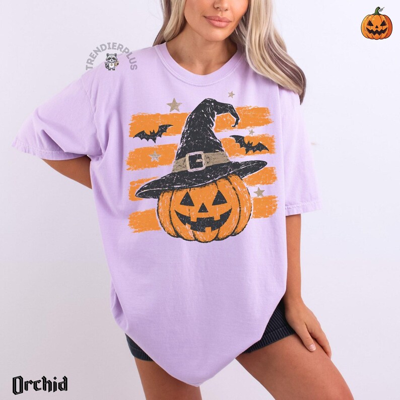 Puede incluir: Una camiseta de color morado claro con un gr&aacute;fico de una calabaza naranja que lleva un sombrero de bruja negro. El dise&ntilde;o incluye trazos de pincel naranjas horizontales, murci&eacute;lagos negros y estrellas peque&ntilde;as. El texto "TRENDIERPLUS" es visible.