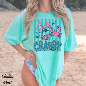 May include: A mint green short-sleeve t-shirt featuring a graphic print with vertical stripes and two pink crabs. The text reads "Cute but a little CRABBY" in pink and blue lettering. The shirt has a loose fit and a crew neckline.