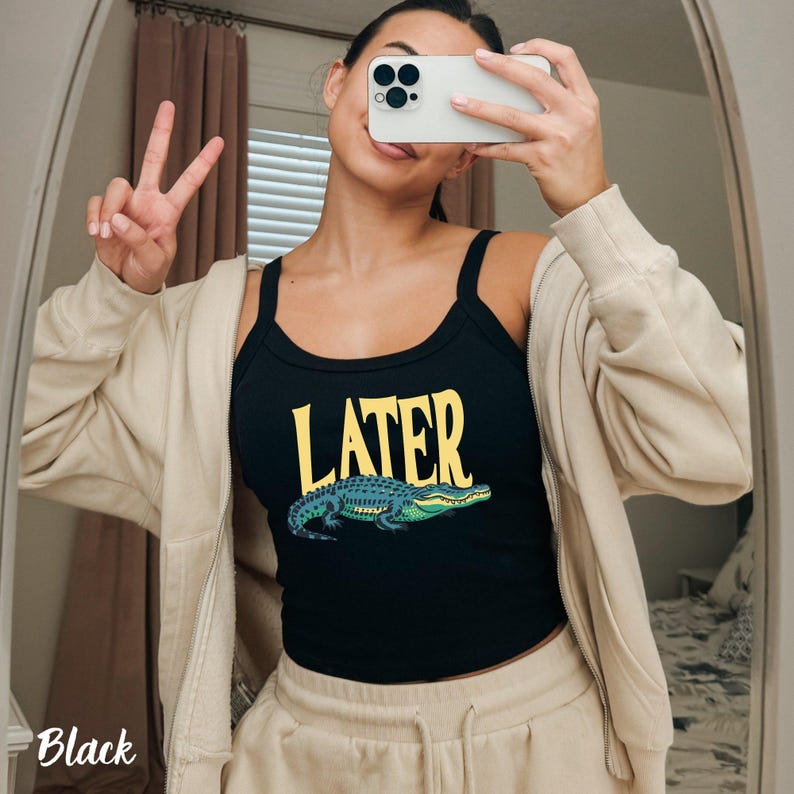 May include: A black tank top featuring the yellow text LATER above a graphic illustration of a green and blue alligator. The tank top has a scoop neckline and thick shoulder straps, worn under a beige zip-up hoodie with matching beige sweatpants.
