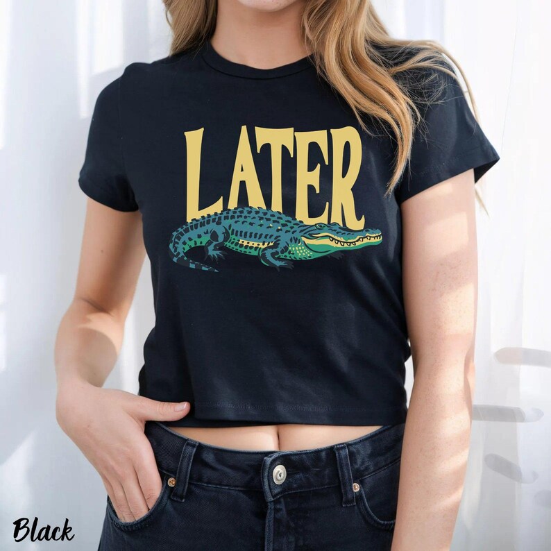 May include: A black short-sleeved t-shirt featuring the word LATER printed in large yellow capital letters above a graphic illustration of a green and blue alligator. The design is centered on the chest of the garment, which has a crew neckline.
