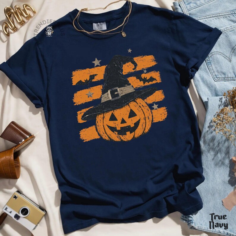 Puede incluir: Una camiseta azul marino con un gr&aacute;fico de una calabaza naranja que lleva un sombrero de bruja negro. El dise&ntilde;o incluye pinceladas horizontales naranjas, peque&ntilde;as estrellas y murci&eacute;lagos. La camiseta tiene cuello redondo y mangas cortas.