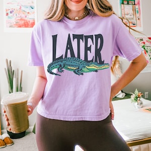 May include: A light purple short-sleeve t-shirt featuring the word LATER in large dark gray capital letters above a graphic illustration of a green and dark blue alligator. The shirt has a crew neckline and a relaxed fit.