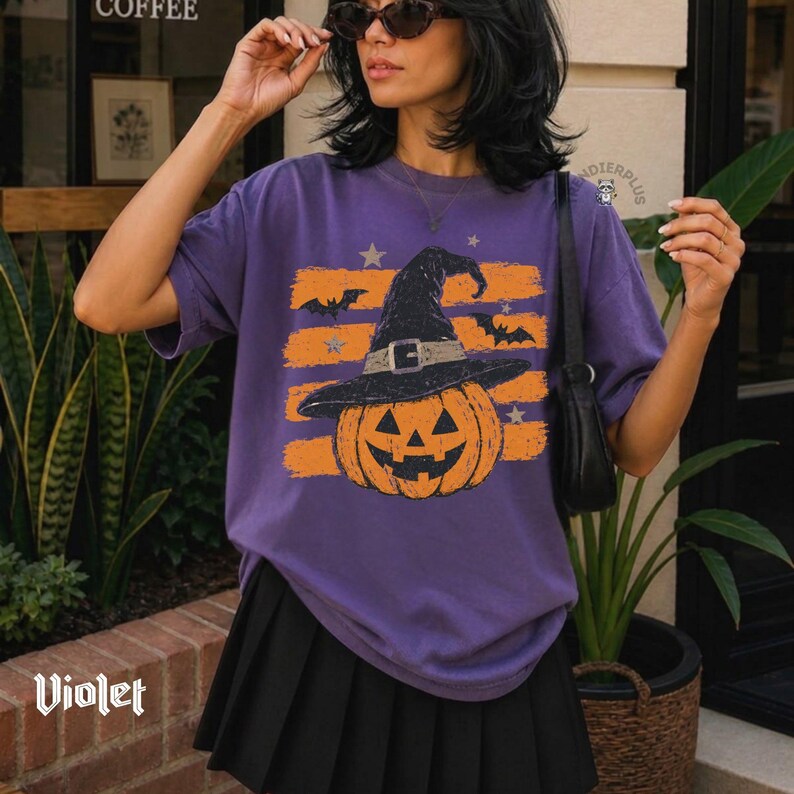 Puede incluir: Una camiseta morada de manga corta con un estampado gr&aacute;fico de una calabaza de Halloween con un sombrero de bruja negro. El dise&ntilde;o incluye rayas horizontales naranjas, estrellas peque&ntilde;as y siluetas de murci&eacute;lagos volando. Falda negra.