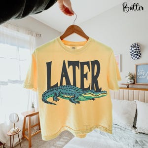 May include: A pale yellow short-sleeve t-shirt featuring the word LATER printed in large black capital letters above a graphic illustration of a green and teal alligator. The shirt is displayed on a wooden hanger against a neutral background.