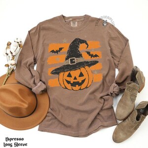 Puede incluir: Una camiseta de manga larga marr&oacute;n con un gr&aacute;fico de una calabaza naranja que lleva un sombrero de bruja negro con hebilla, sobre rayas horizontales naranjas y siluetas de murci&eacute;lagos negros. La camiseta tiene un cuello redondo.
