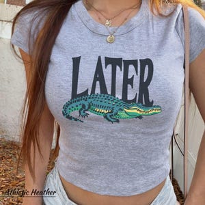 May include: A heather gray short-sleeved t-shirt featuring the word LATER printed in large black capital letters above a graphic illustration of a green and yellow alligator. The shirt has a crew neckline and a fitted silhouette.