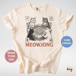 Może przedstawiać: T-shirt z kr&oacute;tkim rękawem w kolorze kości słoniowej z grafiką przedstawiającą cztery koty siedzące przy stole i grające w mahjonga. Pod stołem znajduje się napis MEOWJONG oraz cztery kwadratowe ramki z chińskimi znakami w rogach wzoru.