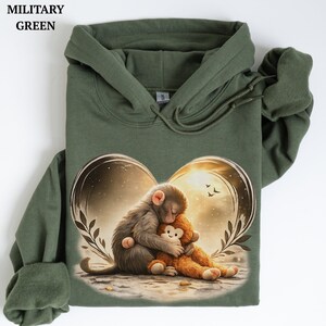 Puede incluir: Una sudadera con capucha de color verde militar que presenta un estampado gr&aacute;fico de un mono abrazando a un mono de peluche. El dise&ntilde;o est&aacute; enmarcado por un contorno en forma de coraz&oacute;n con hojas y un fondo c&aacute;lido y brillante.