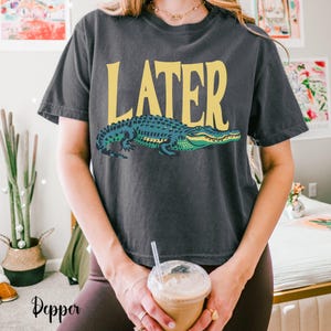May include: A charcoal gray short-sleeve t-shirt featuring the word LATER in large yellow capital letters above a graphic illustration of a green and blue alligator. The shirt has a crew neckline and a relaxed fit, showcasing a screen-printed design.