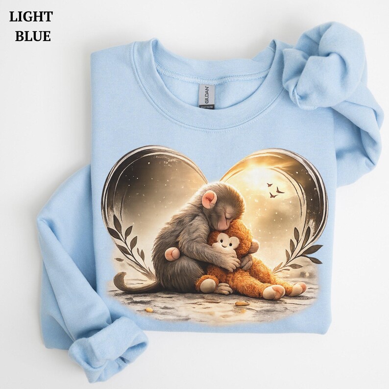 Light blue sweatshirt featuring a graphic of a monkey hugging a teddy bear inside a heart-shaped design. The sweatshirt has long sleeves and a crew neck.