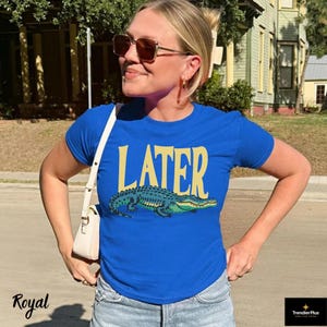 May include: A royal blue short-sleeved t-shirt featuring the word LATER in large yellow letters above a graphic illustration of a green and blue alligator. The shirt is paired with a white shoulder bag and light blue denim jeans.