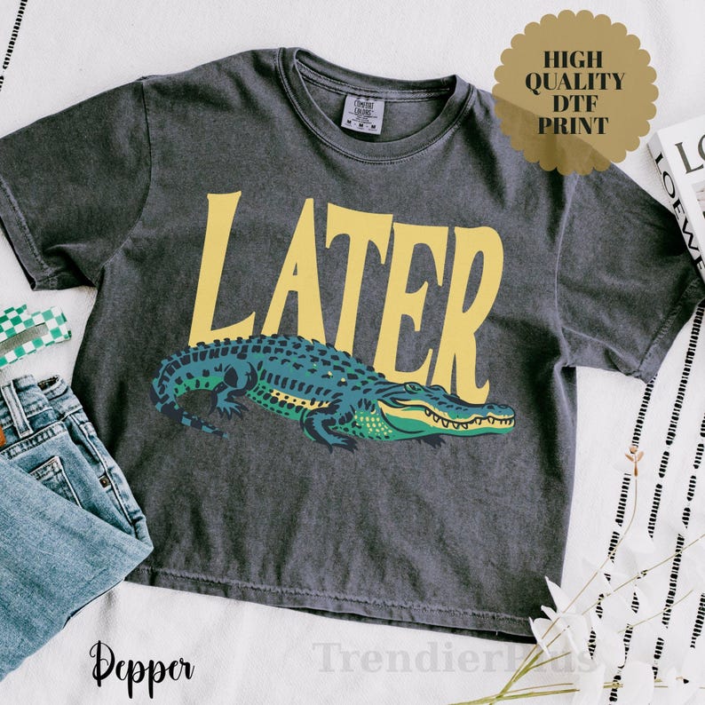 May include: A charcoal gray short-sleeve t-shirt featuring the word LATER in large yellow letters above a graphic of a green and blue alligator. The shirt has a crew neckline and a visible brand tag on the inside of the collar area.