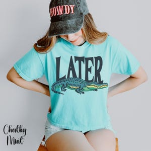 May include: A light turquoise t-shirt featuring a dark graphic of an alligator and the word LATER printed above it. The shirt is paired with a charcoal gray baseball cap that has the word HOWDY embroidered in red and white lettering.