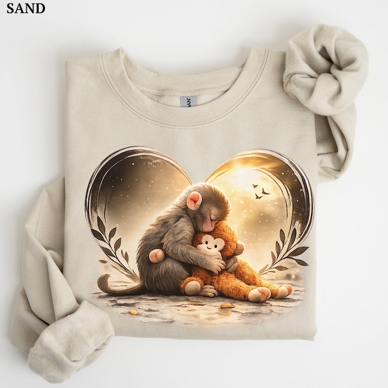 Sand-colored sweatshirt featuring a heart-shaped graphic of a monkey hugging a teddy bear. The illustration has a warm, golden color palette with a sunset background. The sweatshirt has long sleeves and a crew neck.