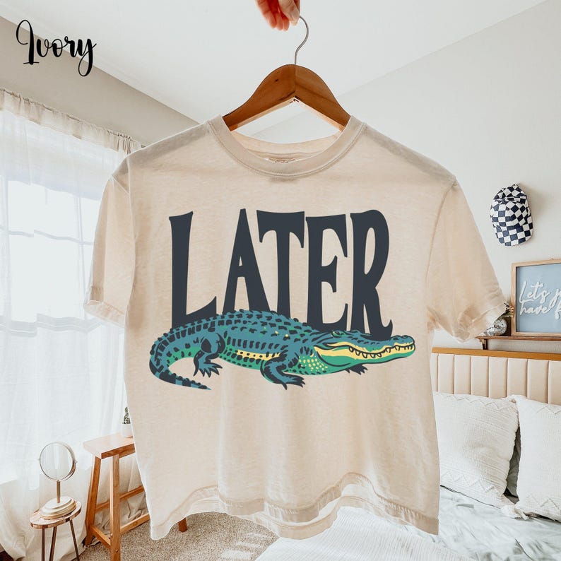 An ivory boxy crop top hanging on a wooden hanger, features a detailed green and teal alligator graphic under the bold black word LATER.
