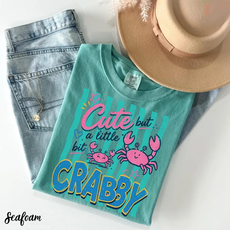 May include: A seafoam green t-shirt featuring a graphic of two pink crabs and the text "Cute but a little bit CRABBY" in pink and blue. The shirt is displayed alongside light blue denim jeans, a tan wide-brimmed hat, and a delicate gold necklace.