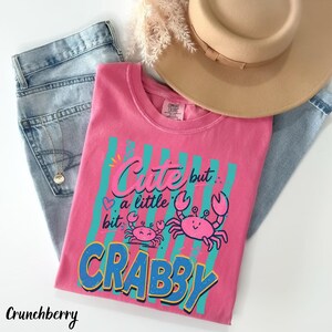 May include: A pink t-shirt featuring a graphic of two pink crabs and the text "Cute but a little bit CRABBY" in blue and pink lettering over a background of vertical teal stripes. The shirt is paired with light blue denim jeans and a tan hat.