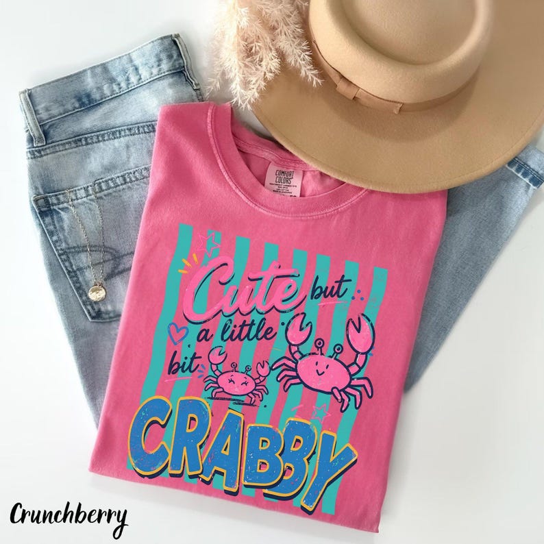 May include: A pink t-shirt featuring a graphic of two pink crabs and the text "Cute but a little bit CRABBY" in blue and pink lettering over a background of vertical teal stripes. The shirt is paired with light blue denim jeans and a tan hat.
