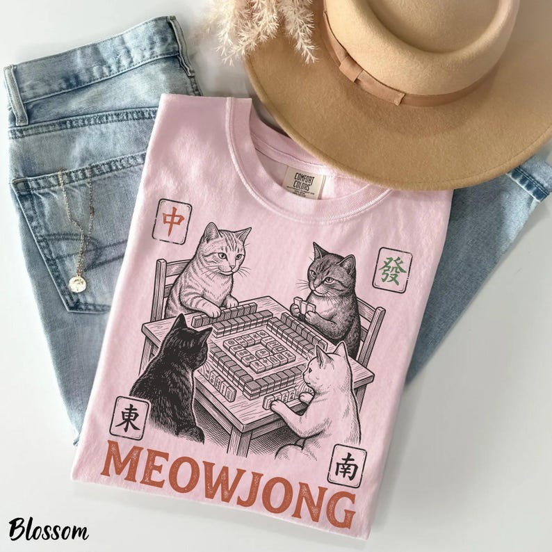 Może przedstawiać: Jasnor&oacute;żowy t-shirt z grafiką przedstawiającą cztery koty grające w mahjonga przy stole. Wz&oacute;r zawiera chińskie znaki w kwadratowych ramkach oraz napis MEOWJONG dużymi brązowymi literami. Koszulka jest położona na jasnoniebieskich dżinsowych spodniach.