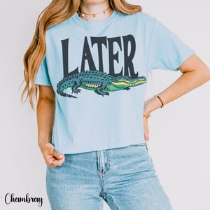 May include: A light blue short-sleeve t-shirt featuring the word LATER printed in large dark charcoal letters above a graphic illustration of a green and yellow alligator. The shirt has a crew neckline and a standard fit.