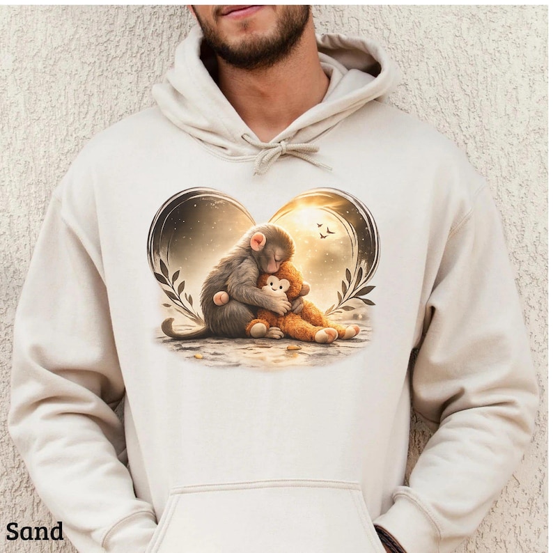 Sand-colored hoodie featuring a heart-shaped design with a gray monkey hugging a brown teddy bear. The design includes a sunset background and leafy accents.