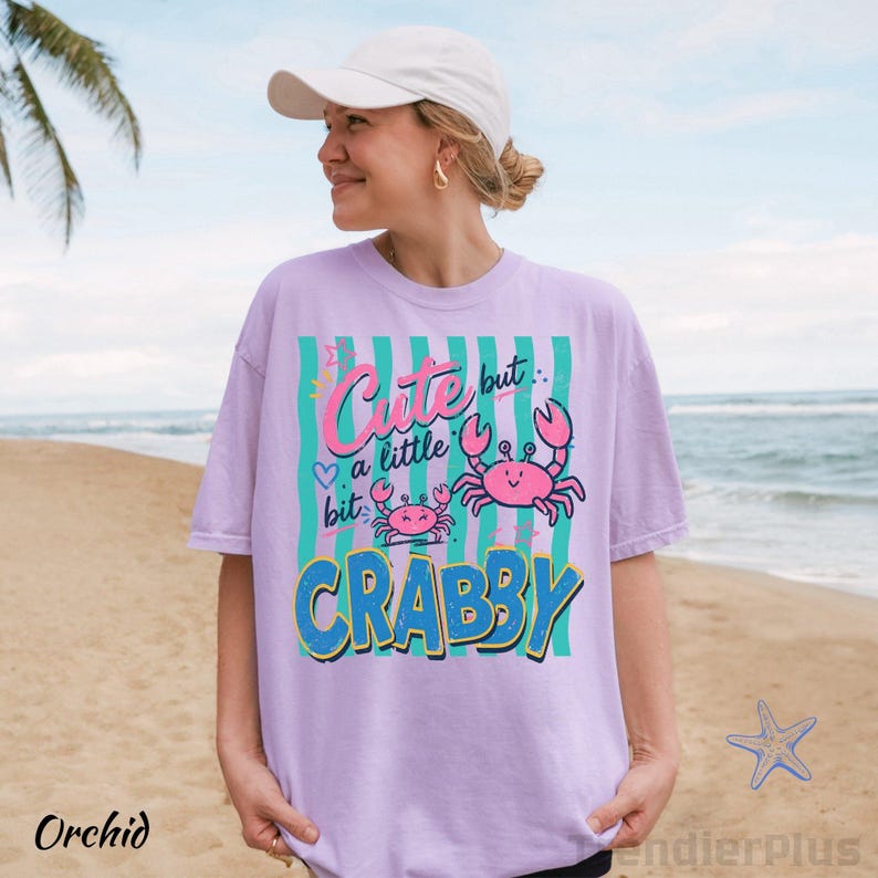 May include: A light purple short-sleeve t-shirt featuring a graphic print with vertical teal stripes, two pink crabs, and the text "Cute but a little bit CRABBY" in pink and blue lettering. The shirt has a crew neckline and a relaxed fit.
