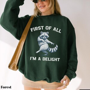 May include: Forest green sweatshirt with the text "FIRST OF ALL I'M A DELIGHT" and a raccoon graphic. The raccoon is depicted in white and gray, with its arms outstretched. The sweatshirt is a solid color.