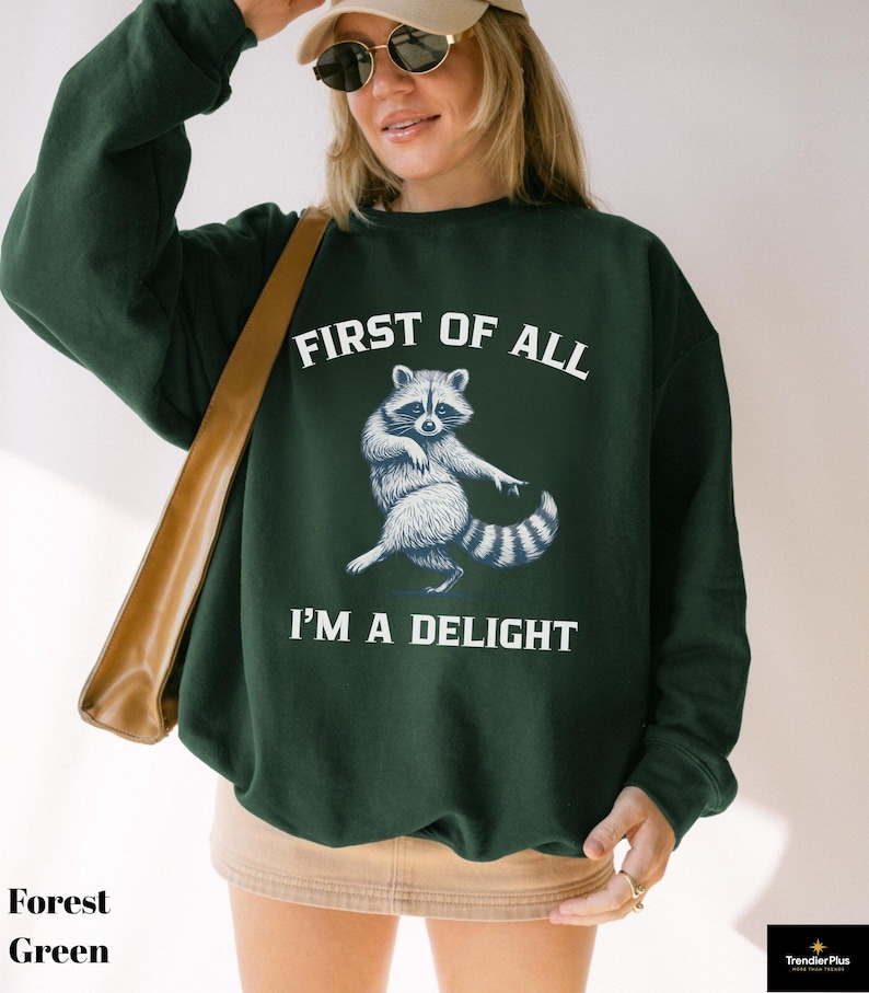 May include: Forest green sweatshirt with the text "FIRST OF ALL I'M A DELIGHT" and a raccoon graphic. The raccoon is depicted in white and gray, with its arms outstretched. The sweatshirt is a solid color.