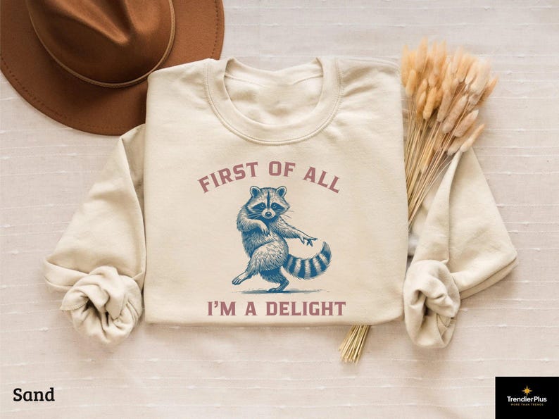 Sand-colored sweatshirt with a raccoon graphic. The raccoon is depicted in blue and white, with the text "FIRST OF ALL I'M A DELIGHT" in a mauve color. The sweatshirt is laid flat with a brown hat and dried flowers.