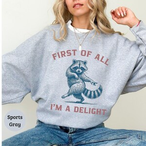 Sports Grey sweatshirt with the text "FIRST OF ALL I'M A DELIGHT" and a dancing raccoon graphic. The raccoon is in shades of blue and gray.