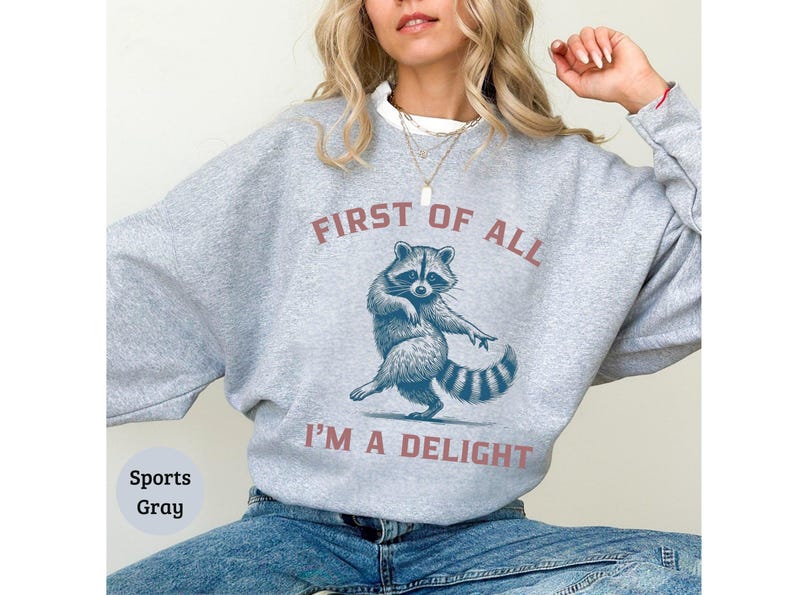 Sports Grey sweatshirt with the text "FIRST OF ALL I'M A DELIGHT" and a dancing raccoon graphic. The raccoon is in shades of blue and gray.