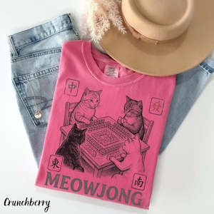 Może przedstawiać: R&oacute;żowy t-shirt z kr&oacute;tkim rękawem z czarną grafiką przedstawiającą cztery koty grające w mahjonga przy stole. Wz&oacute;r zawiera cztery kwadratowe symbole z chińskimi znakami oraz napis MEOWJONG wykonany dużymi szarymi literami na dole.