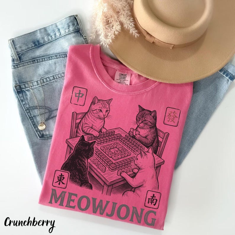 Może przedstawiać: R&oacute;żowy t-shirt z kr&oacute;tkim rękawem z czarną grafiką przedstawiającą cztery koty grające w mahjonga przy stole. Wz&oacute;r zawiera cztery kwadratowe symbole z chińskimi znakami oraz napis MEOWJONG wykonany dużymi szarymi literami na dole.