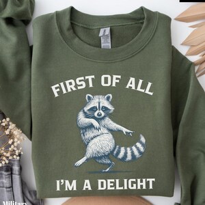 May include: Military green sweatshirt with the text "FIRST OF ALL I'M A DELIGHT" and a dancing raccoon graphic in white. The sweatshirt is folded and has a tag on the collar.