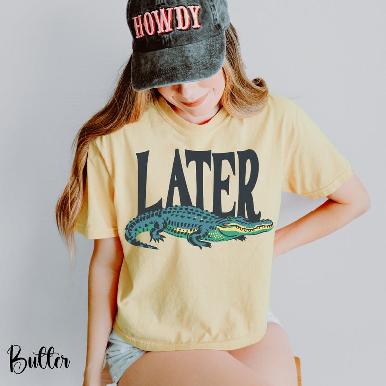 May include: A pale yellow short-sleeve t-shirt featuring the word LATER in large black letters above a graphic of a green and yellow alligator. The t-shirt is paired with a dark gray baseball cap with the word HOWDY embroidered in red letters.