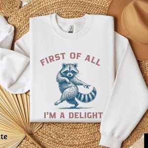 White sweatshirt with the text "FIRST OF ALL I'M A DELIGHT" and a dancing raccoon graphic. The raccoon is in shades of blue and gray.