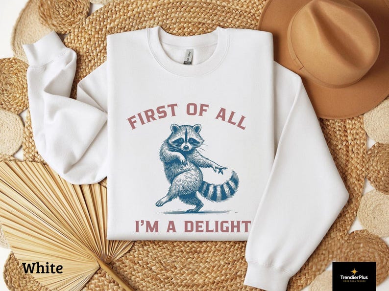 White sweatshirt with the text "FIRST OF ALL I'M A DELIGHT" and a dancing raccoon graphic. The raccoon is in shades of blue and gray.